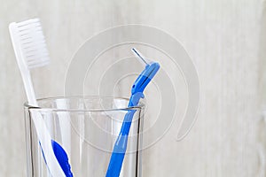 Interdental brush and toothbrush in clean glass on blurred wooden background in bathroom