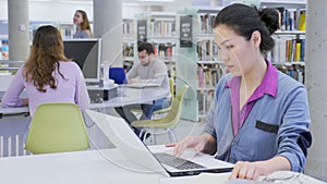 Interested Asian woman sitting at table in library, using laptop, books and taking notes for research. Self-education