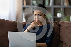 Interested young indian lady recline on sofa using laptop