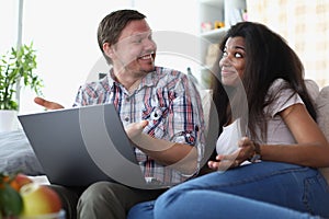 Interethnic family couple laughing and chatting while sitting on sofa at home with laptop.