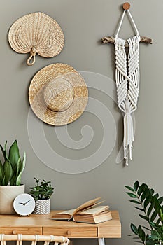 Interior design of boho living room with stylish armchair, wooden desk, house plants, cacti, rattan decoration, macrame and clock.