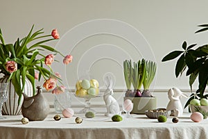 Interior design of easter dining room with colorful easter eggs, white hare sculptures, vase with tulips, plants, lamp, beige wall