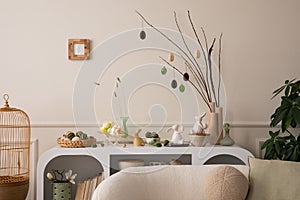 Interior design of easter living room interior with stylish white sideboard, branch with easter eggs, beige bowl, easter bunny,