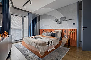 Interior design of elegant bedroom with big orange bed, beige and grey bedclothes, blue curtain, rug, modern lamp, night stand,