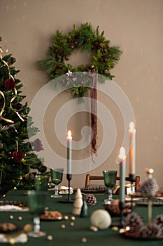 Interior design of warm dining room with table, wreath, christmas tree, green tablecloth, stylish chair, glasses candle with