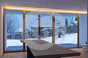 Interior of a dining room, with a large bay window, no one inside and outside there is snow