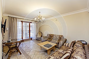 The interior of the living room in beige tones, two sofas and a table. Beige tiles on the floor, a wrought-iron chandelier.