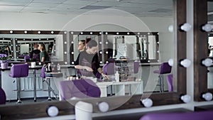 Interior of luxury beauty salon with expert standing and girl sitting near the wall with mirrors. Professional visagiste