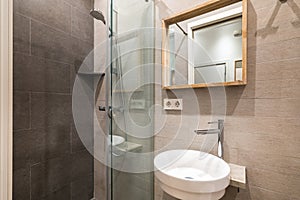 Interior of modern tiled bathroom with white sink, wooden framed mirror and shower zone.