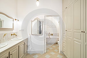 Interior of retro or classic style bathroom decorated in beige color with bath zone, big wardrobe, two sinks and vintage