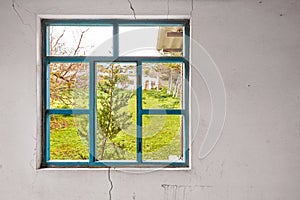 Interior of a ruined house with old, dirty and cracked white wall and a broken window frame with a green meadow field view