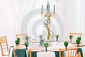 Interior of tent for wedding dinner, ready for guests. Served round banquet table. Golden dishes, green wine glasses and