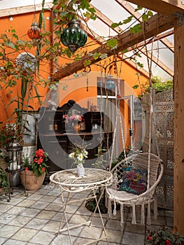 Interior of veranda. Cozy space in patio. A lot of plants. Orange wall. Wooden furniture. Hanging wicker white chair.