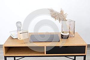 Interior workspace with open blank screen, laptop, vase with dried flowers and statuette in form of head figure.