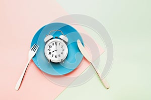 Intermittent fasting concept. White alarm clock on empty blue dish with knife and fork on pink-green background. top view, copy