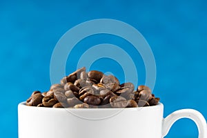 International day of coffee concept. close-up white coffee cup full of coffee beans on blue background.