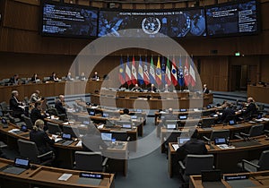 International delegates sit in a modern UN-style conference hall with national flags flying for a global meeting or formal summit. photo