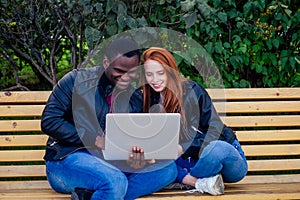 International education concept.dark skinned man wuth redhaired ginger woman looking at laptop and sitting on bench