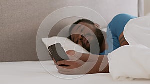 Internet addiction concept. Young indifferent african american guy web surfing on smartphone, lying in bed at home