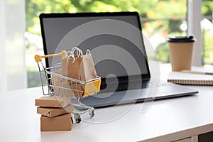 Internet shopping. Small cart with bags and boxes near laptop on table indoors, space for text