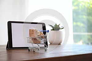 Internet shopping. Small cart with bags and boxes near modern tablet on table indoors, space for text