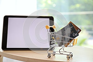 Internet shopping. Small cart with credit cards near modern tablet on table indoors