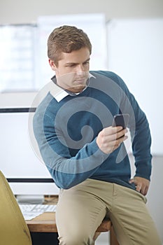 Interoffice communication. a handsome young man using his cellphone while sitting on his desk in an office.