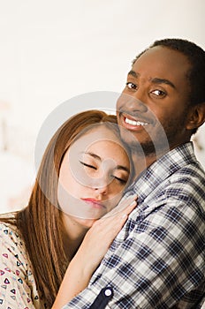 Interracial charming couple wearing casual clothes posing while embracing intimately, she has eyes closed and him