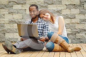 Interracial charming couple wearing casual clothes sitting down on wooden surface lookin at laptop together, in front of