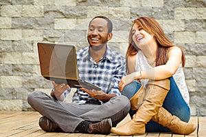 Interracial charming couple wearing casual clothes sitting down on wooden surface lookin at laptop together, in front of