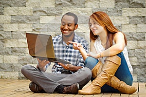 Interracial charming couple wearing casual clothes sitting down on wooden surface lookin at laptop together, in front of