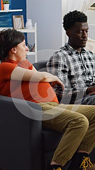 Interracial couple of parents expecting baby sitting on sofa