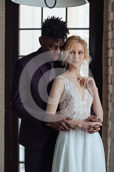 Interracial couple standing in front of a big window. Interracial bridal photoshoot. Caucasian woman getting married