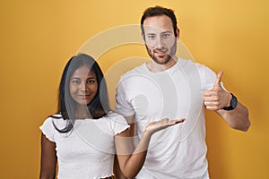 Interracial couple standing over yellow background showing palm hand and doing ok gesture with thumbs up, smiling happy and
