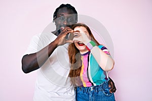 Interracial couple wearing casual clothes doing heart shape with hand and fingers smiling looking through sign