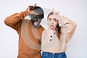 Interracial couple wearing casual clothes making fun of people with fingers on forehead doing loser gesture mocking and insulting