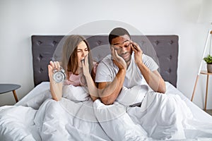 Interracial married couple having problem with waking up in morning, sitting sleepy in bed with alarm clock