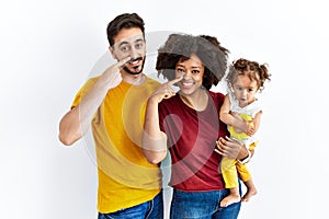 Interracial young family of black mother and hispanic father with daughter pointing with hand finger to face and nose, smiling
