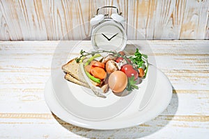 Interval fasting diet concept symbolized by a plate filled to one third with food and an alarm clock on a light wooden background