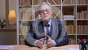 Interview of senior businessman talking calmly and interestingly into camera on book shelves background.