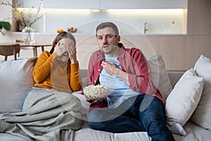 Intrigued man watching spicy film episode with opened mouth, blushing wife closing eyes of confusion