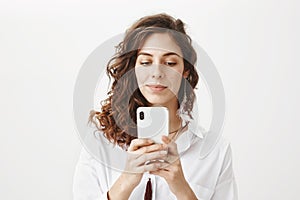 Intrigued and positive sensual caucasian woman with curly hair holding smartphone while messaging or playing games