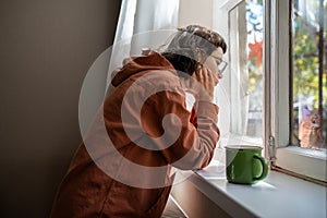 Introverted teen girl standing near window, looking at street indifferently, thinking of life