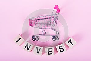 INVEST - word on wooden cubes, on a pink background with a shopping trolley