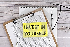 INVEST IN YOURSELF text on yellow sticky on clipboard with chart, wooden background