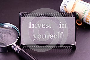 invest in yourself word on the business card next to a roll of money and a magnifying glass on a black background