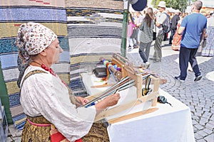 Senior white woman at decorated tent weaving colorful striped belt on hand loom. Irbit fair, Russia