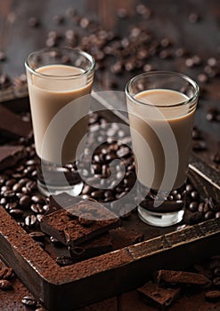Irish cream baileys liqueur in shot glasses in wooden tray with coffee beans and powder with dark chocolate on dark wood
