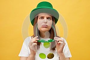 Irish holiday boring party. Sad upset unhappy woman wearing green hat holding clover glasses standing isolated over yellow