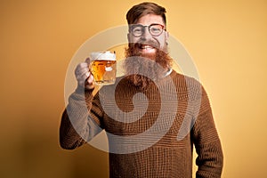 Irish redhead man with beard drinking a glass of refreshing beer over yellow background with a happy face standing and smiling